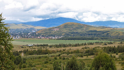 Obraz premium Panoramic view of green mountains and forests with a small town in the valley. Scenic summer landscape with hills, urban skyline and cloudy sky