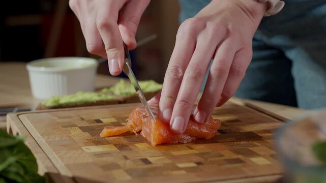 Woman slicing salmon fillet with knife on wooden cutting board with avocado spread on bread and white ramekin in background. Concept of healthy sandwich preparation