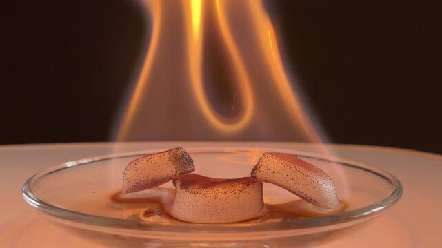 Three solid fuel cubes burn intensely in a glass dish. The dancing orange flames produce a series of quiet cracklings and flickering pops. The fuel melts and darkens as it combusts.