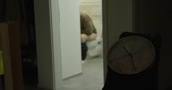Nighttime scene showing a man sitting on the toilet with stomach cramps, viewed through a doorway with a clock in the foreground. Concept of digestive pain, nighttime illness