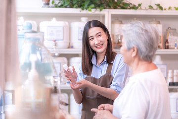Young Asian female entrepreneur showing a reusable bottle to an elderly customer in a modern eco-friendly refill store. Highlighting sustainable shopping.