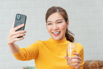 Happy young woman holding a glass of orange water and taking a selfie with her smartphone. Concept of social media, healthy lifestyle, and wellness.