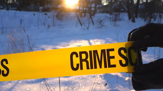 Gloved investigator hand extends caution ribbon across sparkling snow as low sun silhouettes the woodland.