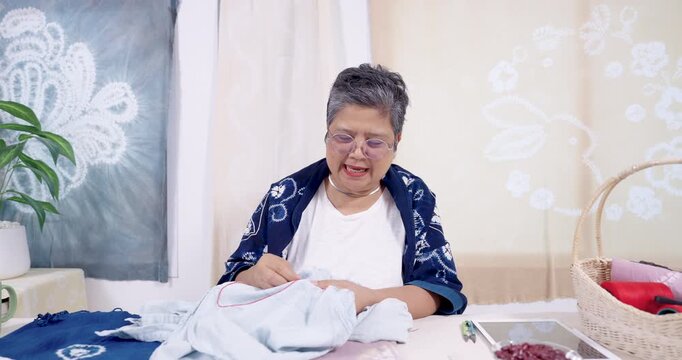 Senior Asian woman artisan smiles, pulls red thread through denim, tightens resist stitch, keeps shibori pattern consistent, talks during live tutorial, handmade textile workflow in home studio.
