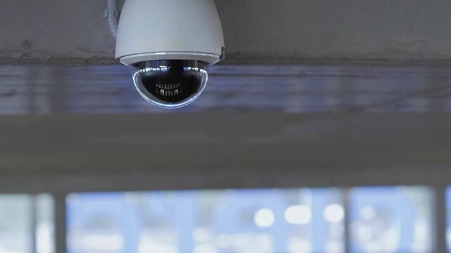 Close-up of a dome security camera on a ceiling in a modern, bright space. Blurred background suggests a spacious interior with windows.