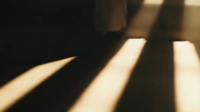 Golden sunbeams stream through a window, casting long, parallel shadows and bright light stripes across a dark, textured floor, creating a warm, abstract pattern