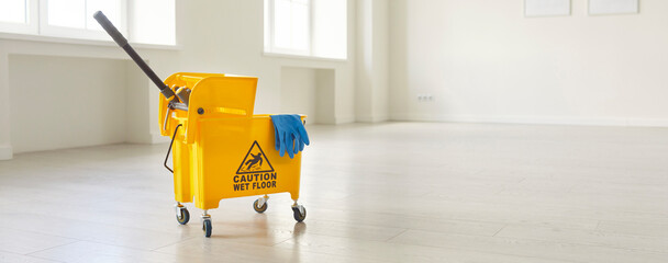 Horizontal photo, banner view of yellow mop bucket with caution sign stands inside empty, spacious room with shiny floors and panoramic windows. Cleanliness, janitorial work, hygiene, facility upkeep © Studio Romantic
