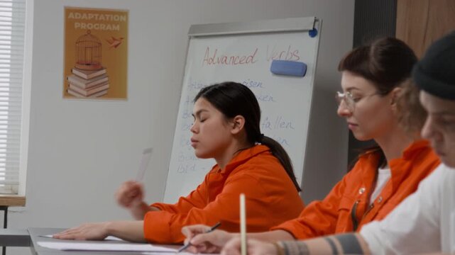 Side view shot of group of young women as female inmates sitting at desks in row and doing tasks during class in prison education program with focus on Latin American woman looking over
