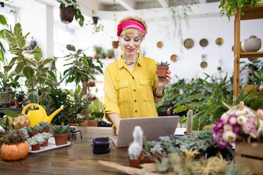 A woman holds a small potted succulent while working on her laptop in a plant shop