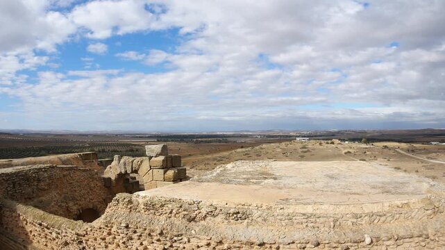 Panorama: Uthina archaeological site Tunisia featuring ancient Roman columns on elevated terrace, wide angle view overlooking countryside beneath dramatic cloudy sky, highlighting classical heritage