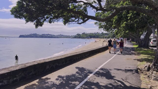 People stroll along a paved path next to Kohi beach in Auckland, New Zealand. The path is lined with trees, providing shade for walkers and beachgoers.