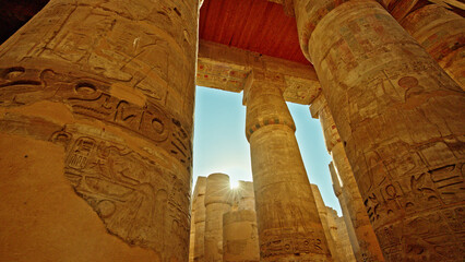 Naklejka premium Ancient stone columns with carved hieroglyphs viewed from below inside Karnak Temple in Luxor Egypt under clear blue sky and warm sunlight