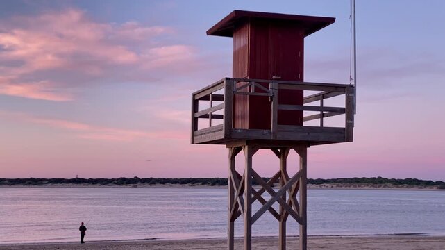 Lifeguard tower on a sandy beach at sunset, showing a gradient sky with a river mouth and boats in the backgrounds