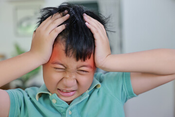 Fototapeta premium Asian boy suffering from severe headache or migraine with red highlight on forehead, child having fever, dizziness, brain tumor symptoms or stress while touching head at home.