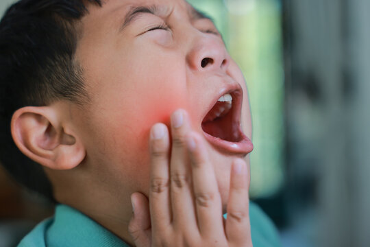 Asian boy suffering from severe toothache with red highlight on cheek, child having dental problem, tooth decay, gum inflammation, sensitive teeth or wisdom tooth pain at home.