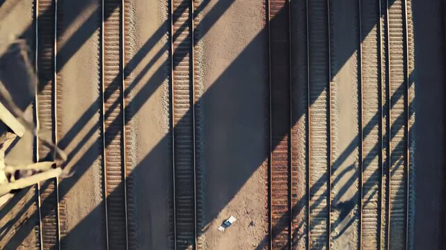 Industrial railway tracks stretching across the frame, with long, dramatic shadows from an overhead steel structure falling across the gravel bed, creating a sense of scale and industrial strength
