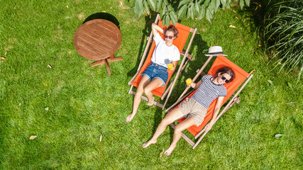 Young girls relax in summer garden in sunbed deckchairs on grass, women friends have fun outdoors in green park on weekend, aerial drone view from above © Iuliia Sokolovska