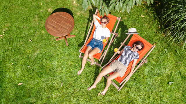 Young girls relax in summer garden in sunbed deckchairs on grass, women friends have fun outdoors in green park on weekend, aerial drone view from above