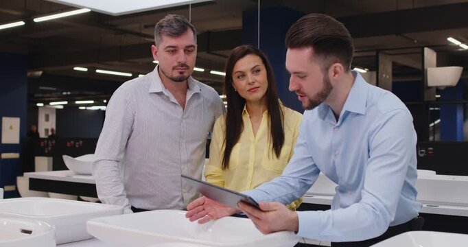 Sale manager helping customers, couple clients to choose bathroom, kitchen sink set, decide type, planning repairs work, interior designer talking to discuss finishing plumbing renovation