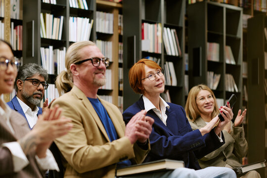 Diverse group of men and women sitting in library attending presentation, holding books and mobile phone, smiling and clapping while looking toward speaker