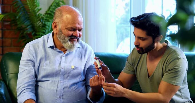 Indian caring young son checking blood sugar level of aged father using digital glucometer while sitting on sofa in modern home, concerned about glucose reading and supporting elderly parent