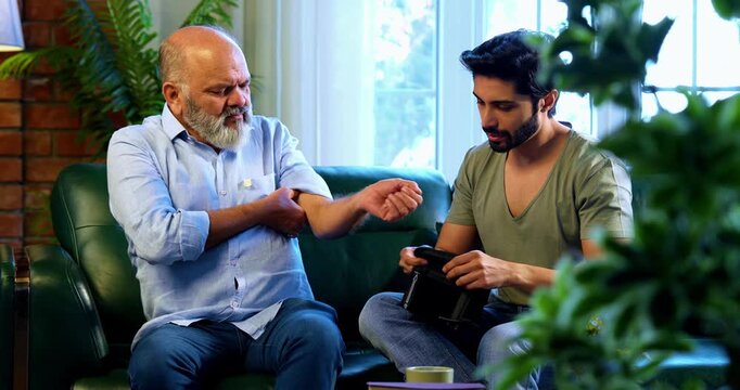 Indian caring young son checking blood pressure of aged father with digital monitor while sitting on sofa in modern home, concerned about health reading and supporting elderly parent with empathy