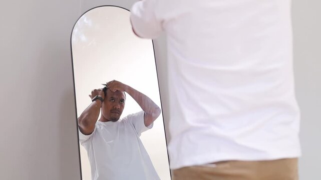 Handsome confident Asian man brushing hair holding comb looking in room mirror, haircare and styling concept.