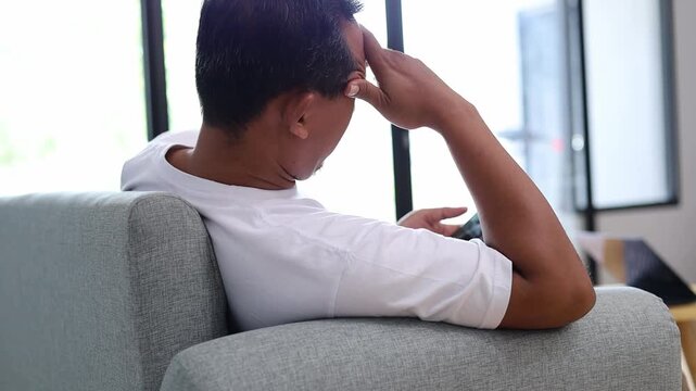 Back view of Bored Asian man sitting on sofa while using smartphone, resting head in hand with tired gesture