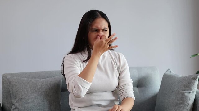 Young asian woman sitting on the sofa at home smelling something stinky and disgusting, intolerable smell, holding breath with fingers on nose. bad smell