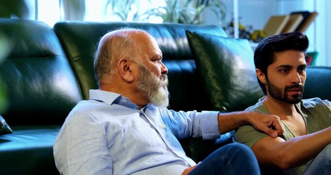 Indian young son and aged father enjoying coffee together and engaged in light hearted conversation while sitting on carpet in airy living room, sharing laughter, bonding and quality time at home