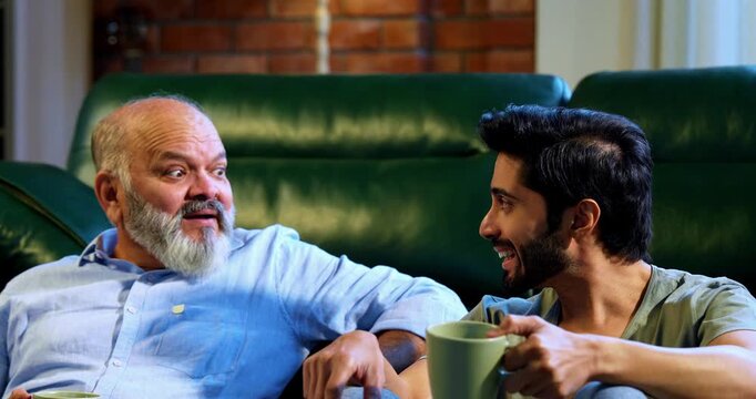 Indian young son and aged father enjoying coffee together and engaged in light hearted conversation while sitting on carpet in airy living room, sharing laughter, bonding and quality time at home