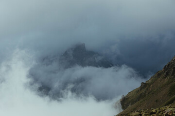 Fog and clouds surround a mountain peak, creating a dramatic atmosphere in a high-altitude area. scene shows rocky terrain and distant rock formations