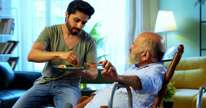 Indian young son feeding senior father chapati with sabzi from plate while he relaxes on chair, sharing heartfelt conversation and affection in modern home living room during homely mealtime