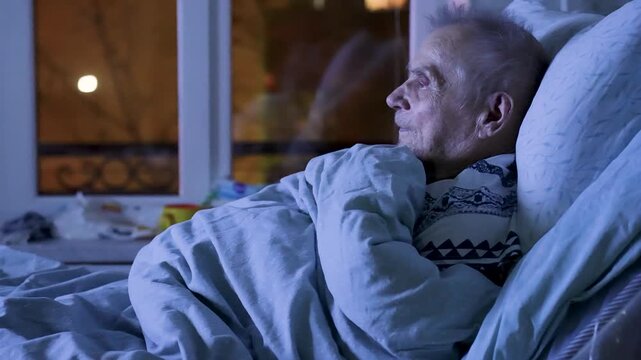 Senior Man Lying in Bed Watching Television at Night. Elderly man resting in bed in a dimly lit room, looking at a bright TV screen. Concept of senior lifestyle, aging, loneliness