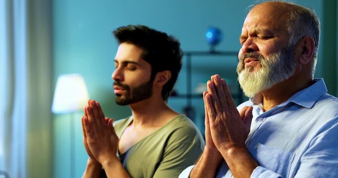Indian senior father and young son praying with folded hands and chanting mantra as daily devotional ritual practice in modern Hindu home living room, expressing faith, spirituality and family unity