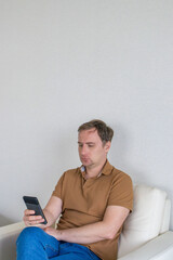 Fototapeta premium Adult man sitting in armchair using smartphone at home or hotel room. Online communication, social media, mobile banking and digital lifestyle concept.