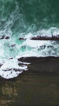 Dynamic drone capture of turquoise waves smashing into layered rock formations at Tanjung Layar, Sawarna Beach, Indonesia. Detailed water splashes and textures presented in striking Vertical format.