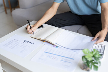 Asian Man Taking Notes on Finances, Budget, Household Expenses, and Tax Calculations in Notebook at Home.