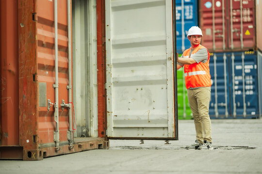 A logistics worker in safety vest and white hard hat pulls open a heavy shipping container door at a cargo yard. He checks the interior before closing it again, illustrating daily warehouse operations