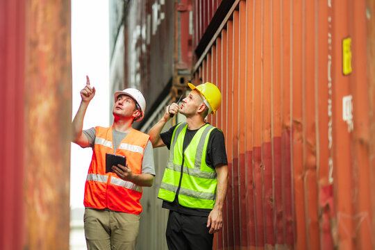 Two cargo inspectors observe upper container levels and communicate via handheld radio. They conduct operational safety audit in maritime logistics hub.