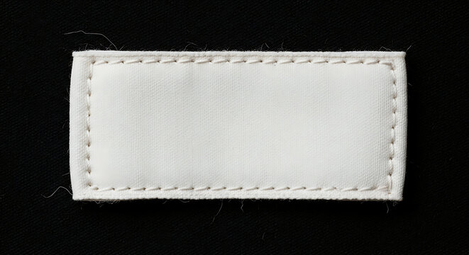 Blank white rectangular fabric clothing label sewn on black textile background, close-up