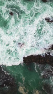High-angle drone footage of thick sea foam spreading across rocky reef surfaces at Tanjung Layar, Sawarna Beach, Indonesia. Dynamic coastal scene recorded in immersive Vertical composition.
