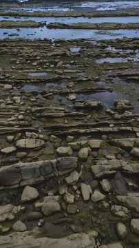 Top-down drone footage showcasing intricate rock layers and shallow water pools at Tanjung Layar Beach, Sawarna, Indonesia. Earthy tones and patterns framed in clean Vertical format.