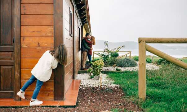 Back view of little girl spying couple in love kissing next to wooden house