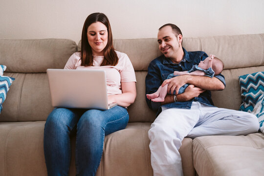 Father carrying sleeping daughter while looking at woman using laptop on sofa