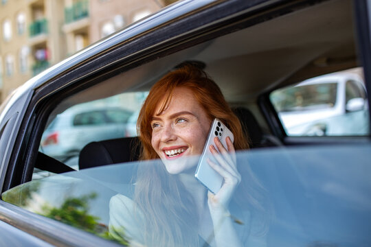 Redhead woman talking on phone in car. Happy female passenger with freckles using smartphone. Communication and transportation concept. Beautiful ginger girl smiling on a call in taxi back seat.