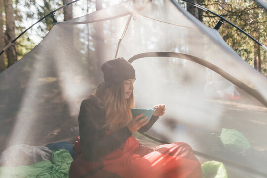 Young woman sitting in tent, eating breakfast in the forest
