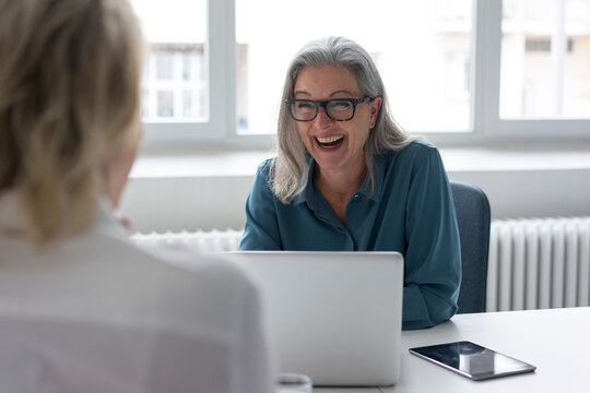 Laughing mature businesswoman talking to young businesswoman at desk in office
