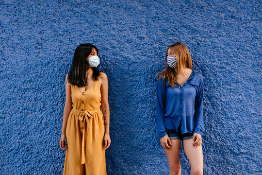 Female friends wearing masks looking at each other while maintaining distance against blue wall in city