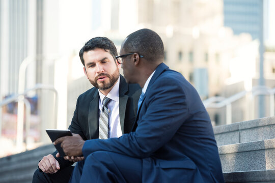 Two businessmen sitting on stairs talking and sharing tablet